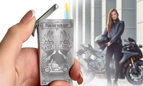 Engraved stainless steel lighter held in a hand, featuring a motorcycle design, with a female biker standing beside her motorcycle in the background