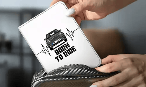Person holder hvit lommebok med svart bil og hjertestopp-design, BORN TO RIDE-tekst, karbonfiberveske i bakgrunnen