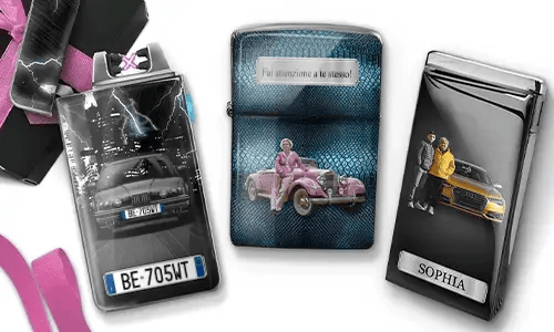 Drei Metallfeuerzeuge mit personalisierten Designs. Das linke Feuerzeug zeigt eine Stadtsilhouette und ein Paar neben einem Auto mit der Aufschrift „BESTE OMA DER WELT“. Das mittlere Feuerzeug stellt ein rosafarbenes Oldtimer-Auto dar, darüber steht „HAB DICH LIEB OMA“. Das rechte Feuerzeug zeigt ein blaues Auto und ein Kennzeichen. Rosafarbene Schleifen verleihen dem Ganzen einen Geschenkcharakter.