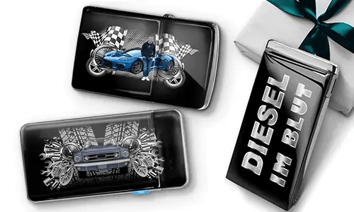 Two sleek black lighters featuring car themes, one with a blue car and checkered flags, another with a red car and gears. A third lighter reads 'DIESEL IM BLUT'. Gift-wrapped in the background.