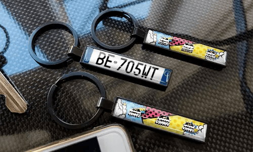 Comic-style license plate keychains displayed on a carbon-textured surface with colorful car designs