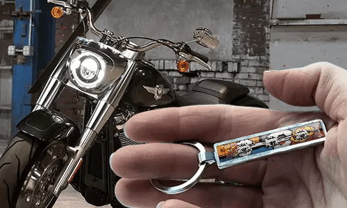 Kovový komiksový přívěsek na klíče s ilustrací motocyklu, držený v ruce vedle motocyklu