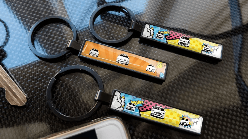 Nøgleringe med nummerplade i tegneseriestil med farverige bildesigns, vist på en kulfibertekstureret overflade